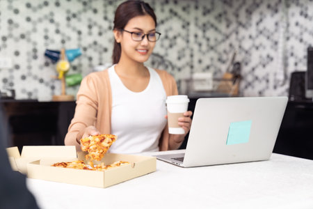 Asian woman work from home in the kitchen and eating delivery pizza take away food and take out coffee while city lockdown from coronavirus covid-19 pandemic. New Normal lifestyle working from home.の写真素材