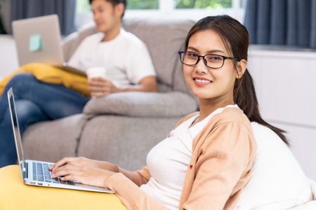Asian working woman use computer notebook laptop work from home in living room with social distance to her couple in background while city lockdown. Work at home concept.の写真素材