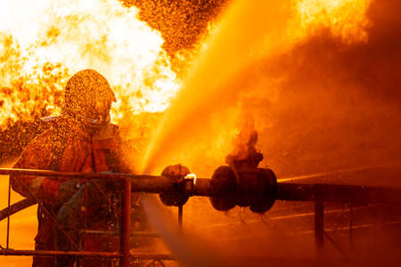 Firefighter using water fog type fire extinguisher to fighting with the fire flame from oil pipeline leak and explosion on oil rig and natural gas station. Firefighter and industrial safety concept.の写真素材