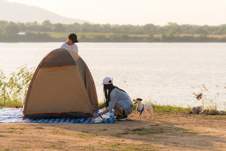 Young adult Asian couple pitch and set up tent for camping around the lake. Camping trip with dog outdoor activity campsite concept.の写真素材