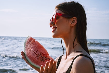 cheerful woman in black swimsuit eating watermelon on the beach