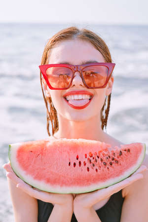 cheerful woman in sunglasses eating watermelon by the oceanの写真素材