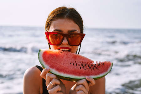 cheerful woman in sunglasses eating watermelon by the oceanの写真素材