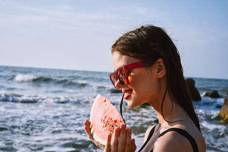 cheerful woman in black swimsuit eating watermelon on the beachの写真素材