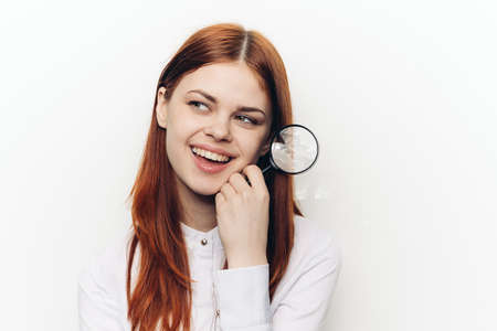 cheerful red-haired woman holding a magnifying glass near her faceの写真素材