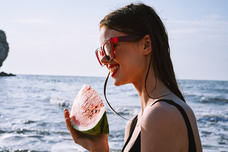 cheerful woman in black swimsuit eating watermelon on the beachの写真素材