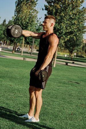 athletic man in black t-shirt with dumbbells in the park training