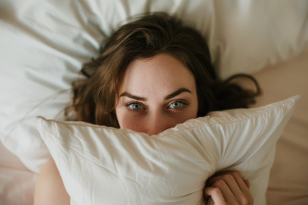 Woman peeking out from under a pillow in bed with wide open eyes, concept of insomnia and stressの素材
