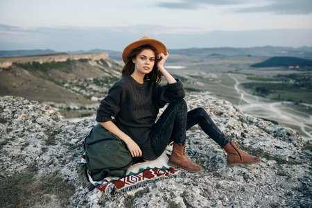 Adventurous woman with backpack and hat sitting on rock at mountain summitの写真素材