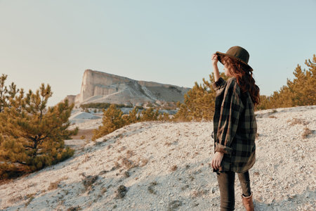 Woman in plaid shirt and hat standing on hill, admiring mountain view on sunny dayの写真素材