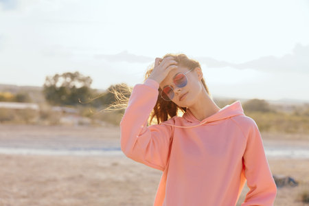 Confident woman in pink hoodie and sunglasses standing in desert with hands on head under bright sunの写真素材