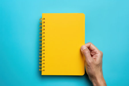 Bright yellow notebook held by a hand against a solid turquoise backgroundの素材