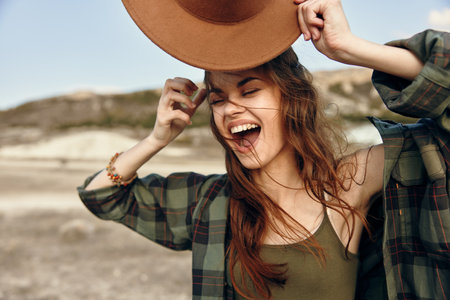 Joyful woman holding hat in field with mountains, laughter and happiness in beautiful natural landscapeの写真素材