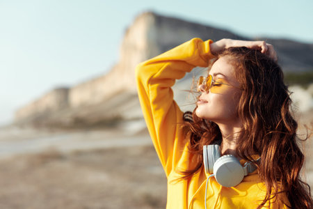 Serene young woman in yellow sweater and sunglasses enjoying music with headphones against mountain backdropの写真素材