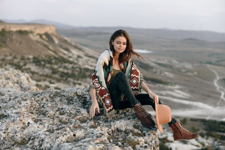 Serene woman in cozy sweater and boots enjoys mountain view from rocky perch on a crisp dayの写真素材