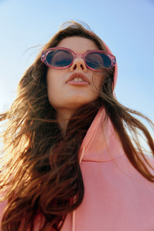 Stylish woman in pink hoodie and sunglasses posing against vibrant blue skyの写真素材