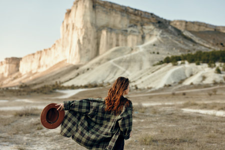Stylish woman in plaid coat and hat gazes at majestic mountain backdrop on a sunny dayの写真素材