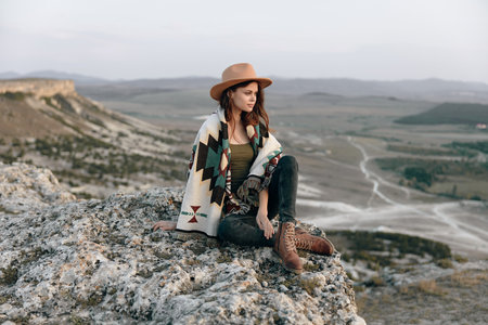 Woman in hat and sweater sitting on mountain rock edge with boots, looking out into the distanceの写真素材