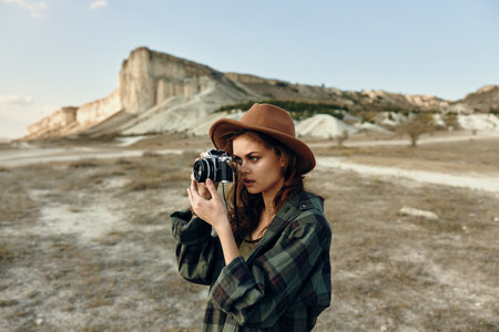 Stylish woman in plaid shirt and hat capturing a moment with vintage camera outdoorsの写真素材