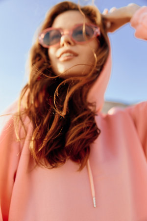 Stylish woman in pink hoodie and sunglasses striking a pose against vibrant blue sky backgroundの写真素材
