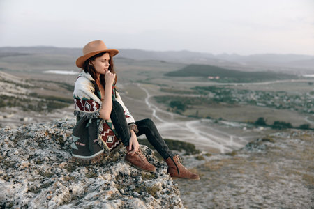 Serene woman in hat and blanket sitting on rock, gazing over picturesque valley landscape