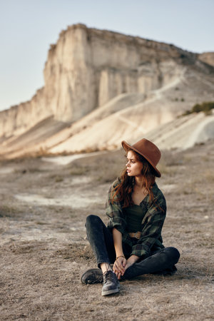 Woman in stylish hat enjoys scenic mountain view while sitting on groundの写真素材