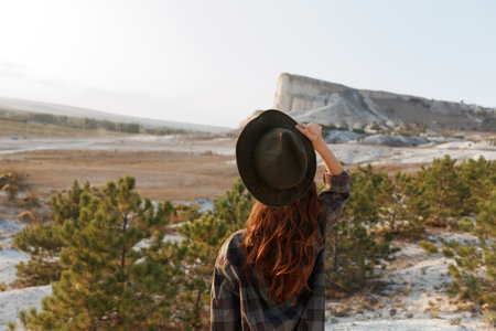 Serene woman in plaid shirt and hat standing on hill with majestic mountain backdropの写真素材