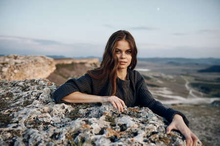 Confident woman sitting on rocky ledge, hands on hips, overlooking majestic mountain landscapeの写真素材