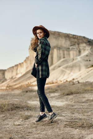 Stylish woman in plaid shirt and hat stands confidently in desert landscape with majestic mountains...
