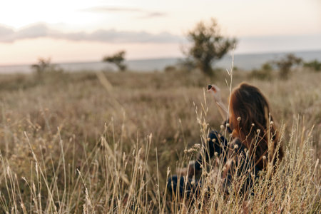 Woman sitting in tall grass at sunset, holding cell phoneの写真素材
