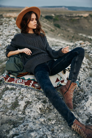 Serene woman in hat and sweater sitting on rock with feet in air enjoying peaceful nature moment