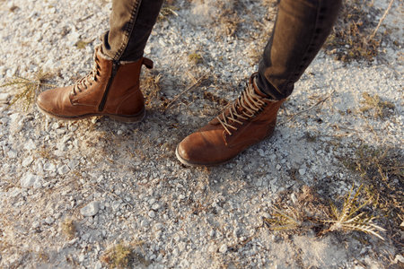 Rugged brown boots standing on a rustic dirt road with grass and rocks in the backgroundの写真素材