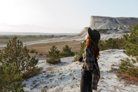 Woman in plaid shirt and hat standing on hill admiring scenic valley view on sunny dayの写真素材