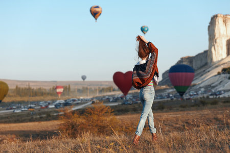 Woman in denim jeans and backpack standing in front of a field with hot air balloons in the backgroundの写真素材