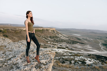 adventurous young woman standing on cliff overlooking valley in green tank top and black jeansの写真素材