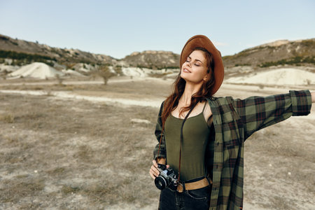 Woman photographer in plaid shirt and hat capturing scenic mountain landscape in open fieldの写真素材