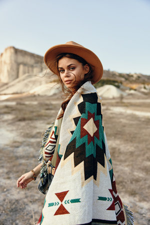 Woman in blanket and hat standing in desert with mountain backdrop on a sunny day