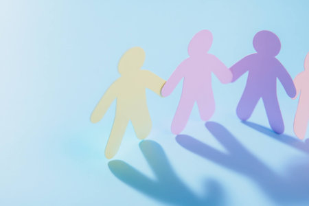 Colorful paper cutouts of people holding hands against a pastel blue background, symbolizing unity, friendship, and diversity in a creative, cheerful concept.の素材