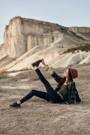 Woman in hat relaxing on ground in front of majestic mountain with legs up in air