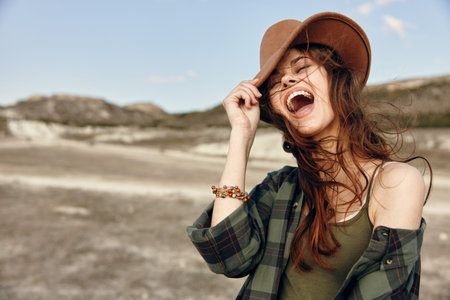 Joyful woman in plaid shirt and brown hat laughing in expansive field on sunny day