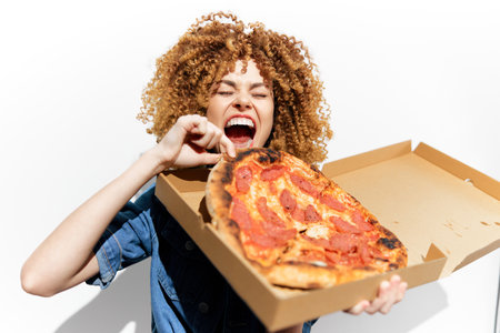Young woman enjoying a large slice of pepperoni pizza from a pizza box, showcasing excitement and joy with curly hair against a bright white backgroundの写真素材