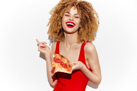 Happy young woman with curly hair enjoying a slice of pizza, wearing a red top, vibrant colors, health concept, playful mood, against a clean white backgroundの写真素材