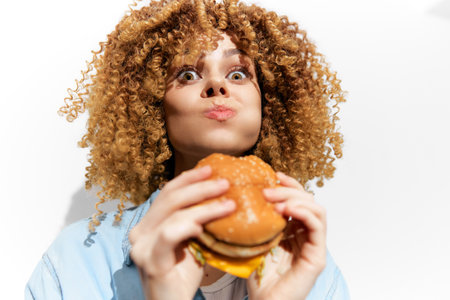 Cheerful young woman with curly hair enjoying a delicious hamburger, showcasing excitement and indulgence against a clean white backgroundの写真素材