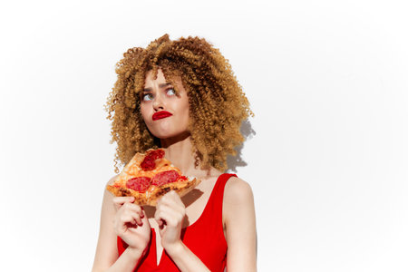 Young woman with curly hair holding a slice of pepperoni pizza, wearing a red dress, looking playfully pensive, ideal for concepts of indulgence and lifestyleの写真素材