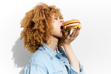 Young woman with curly hair enjoying a hamburger against a white background, embodying the concept of indulgence and comfort food in a vibrant, modern styleの写真素材