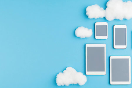 Modern digital devices on a blue background with fluffy clouds, showcasing smartphones and tablets in a playful, whimsical conceptの素材
