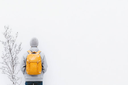 A solitary figure wearing a grey sweatshirt and a knitted hat stands against a minimalistic white wall, emphasizing winter solitude with a yellow backpackの素材