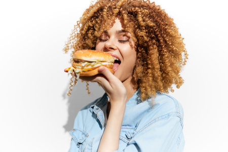 Happy young woman with curly hair enjoying a delicious burger against a bright white background, embodying the joy of indulgenceの写真素材