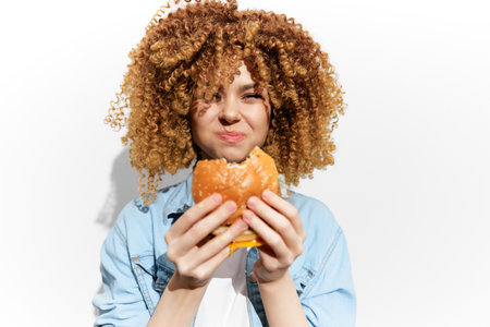 Young woman with curly hair joyfully holding a burger, showcasing a vibrant expression of satisfaction against a white backgroundの写真素材