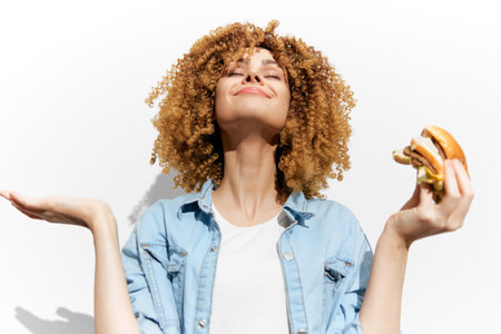 Happy woman enjoying a delicious burger, showcasing a carefree attitude and vibrant curly hair against a bright white backgroundの写真素材